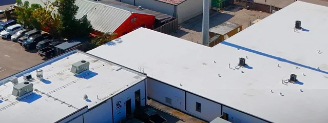 Aerial view of flat, white-roofed commercial buildings with air conditioning units on top, surrounded by parked cars and trees in The Woodlands industrial area—quality Commercial Roofing by Noble Roofing stands out.
