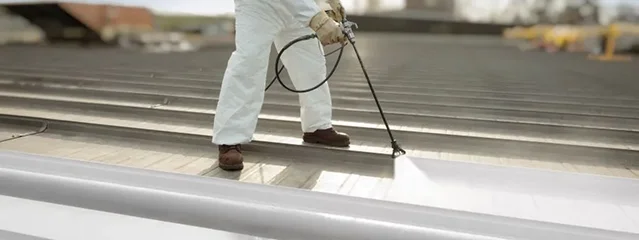 A person in protective clothing sprays a white coating onto a metal rooftop using a spray gun, likely applying a protective or reflective layer as part of commercial roofing services in The Woodlands.