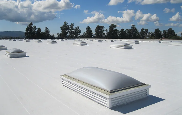 A wide, flat white rooftop with multiple dome-shaped skylights and rectangular vents showcases expert commercial roofing under a blue sky with scattered clouds and distant trees on the horizon in The Woodlands.